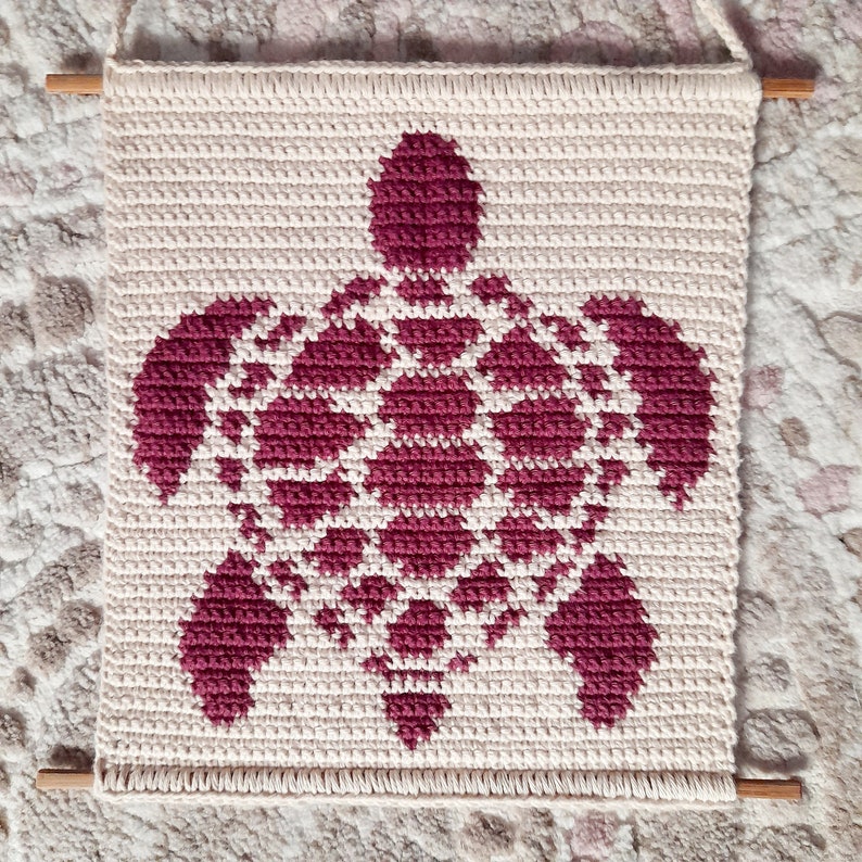 May include: A crocheted wall hanging featuring a burgundy sea turtle design on a cream background. The hanging is framed with wooden dowels.
