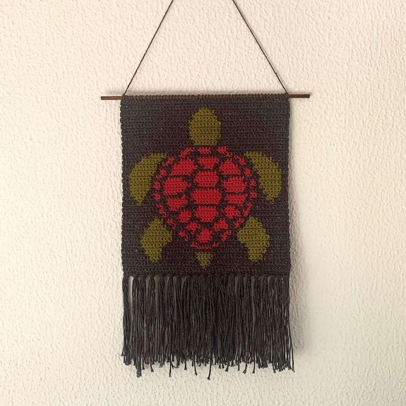 May include: A crocheted wall hanging featuring a red and green turtle design with a black fringe.