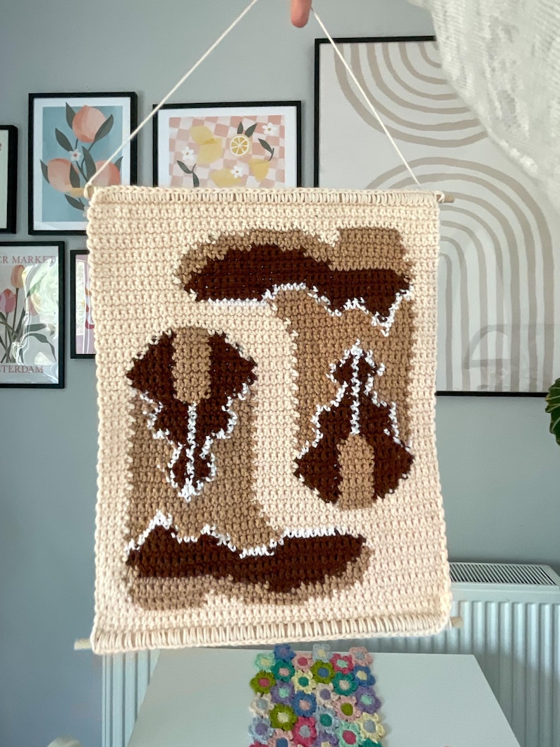 Puede incluir: Un tapiz de pared de ganchillo beige con un dise&ntilde;o de bota marr&oacute;n y beige. La obra de arte est&aacute; suspendida por un pasador de madera y una cuerda blanca. El fondo incluye arte enmarcado y una mesa blanca.