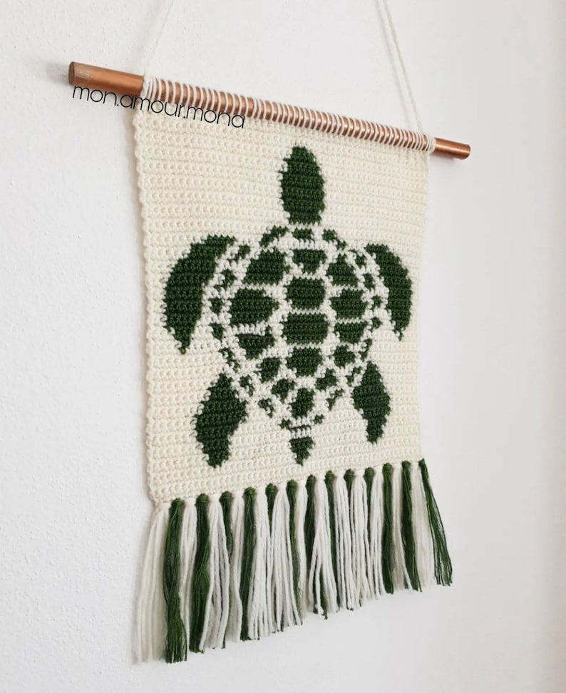May include: A crocheted wall hanging featuring a green turtle design with white fringe. The hanging is attached to a wooden dowel rod.