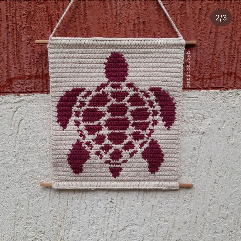 May include: A crocheted wall hanging featuring a burgundy turtle design on a cream background. The hanging is attached to a wall with wooden dowels. The text "@stitch.byh" is visible in the bottom right corner.
