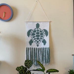 May include: A white crocheted wall hanging with a green sea turtle design and a fringe border.