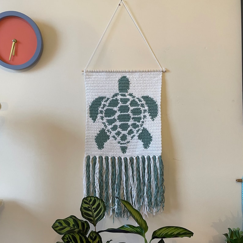 May include: A white crocheted wall hanging with a green sea turtle design and a fringe border.