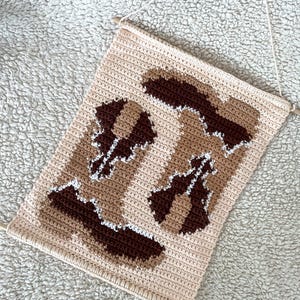 Puede incluir: Un tapiz de pared de ganchillo beige con un dise&ntilde;o geom&eacute;trico en tonos marrones y blancos. La pieza rectangular est&aacute; suspendida por una clavija de madera y una cuerda para colgar. El dise&ntilde;o presenta formas abstractas.