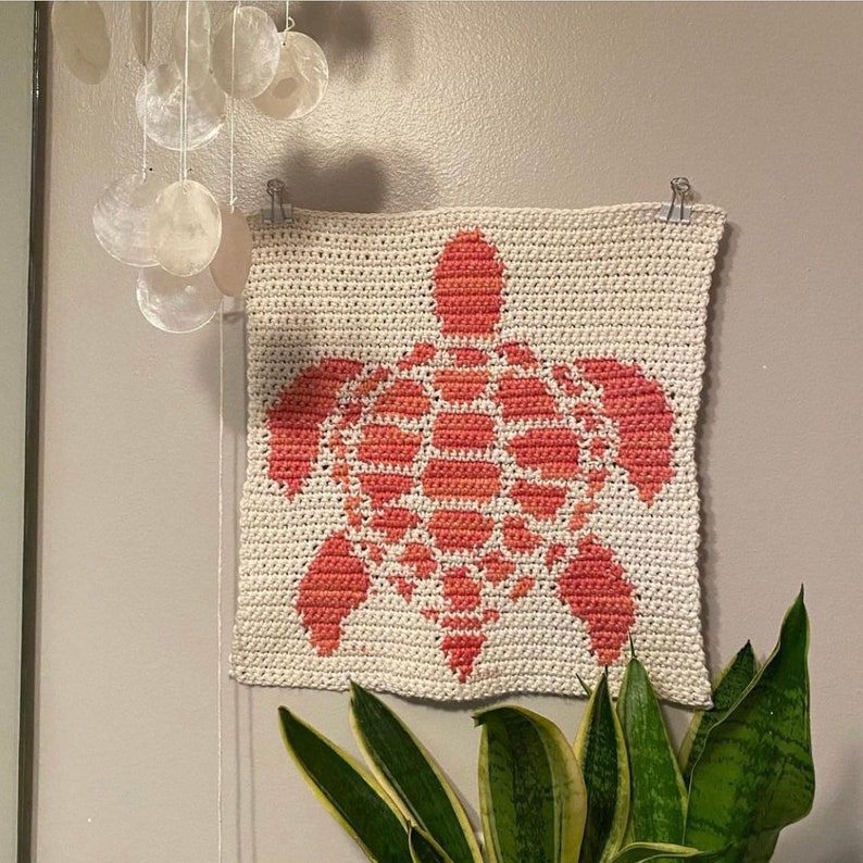 May include: A crocheted wall hanging featuring a coral-coloured sea turtle design on a white background.