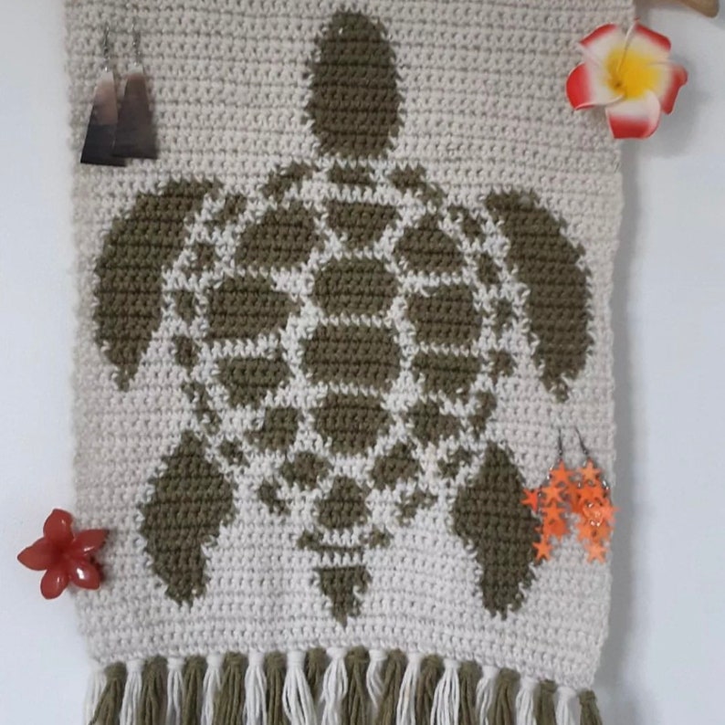 May include: A crocheted wall hanging featuring a green turtle design with white accents. The hanging has a fringe border and is made with a beige yarn.