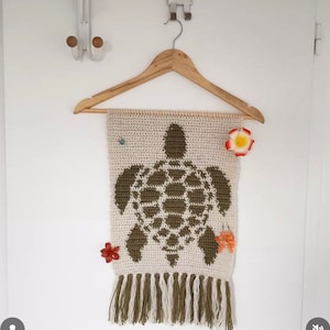 May include: A crocheted wall hanging featuring a green turtle design with a white background. The hanging has a fringe border and is displayed on a wooden hanger.