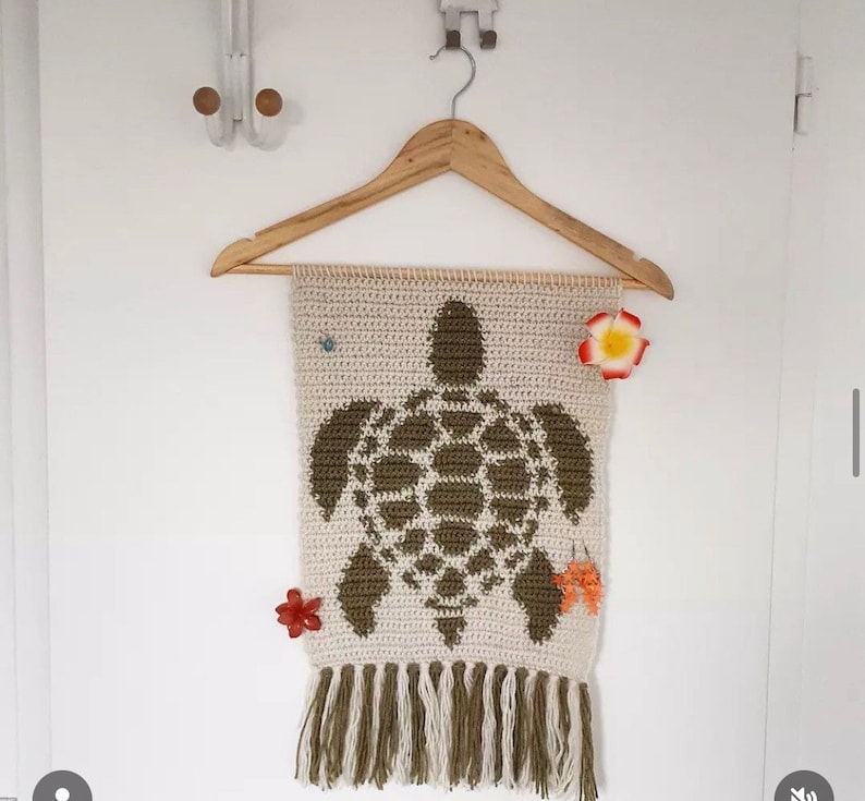 May include: A crocheted wall hanging featuring a green turtle design with a white background. The hanging has a fringe border and is displayed on a wooden hanger.