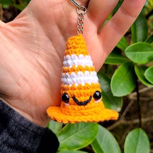 May include: A crocheted orange and white traffic cone keychain with a black smiling face and black eyes.