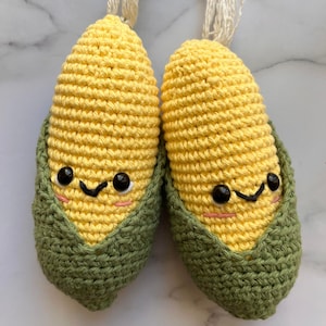 May include: Two crocheted corn on the cob toys with green husks and yellow kernels. Each corn has a smiling face with black eyes and a pink mouth.