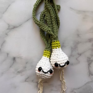 May include: Two crocheted green onions with white bulbs and yellow accents. The onions have black eyes and a smiling mouth. The onions are on a white marble background.