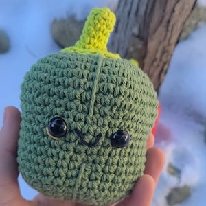 May include: A green crocheted plush toy shaped like a bell pepper with a yellow stem and black eyes. The toy has a stitched-on smile.