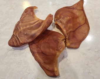 May include: Three brown pig ears, a popular dog chew treat, are arranged on a white countertop.