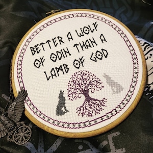 May include: A white embroidery hoop with a black and purple cross-stitch design. The design features a tree with roots, two wolves, and the text "Better a wolf of Odin than a lamb of God."