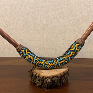 Könnte beinhalten: Eine gebogene Pfeife mit Holzstielen und einem perlenbesetzten Körper. Der Körper ist mit einem geometrischen Muster aus türkisfarbenen, gelben und schwarzen Perlen verziert. Die Pfeife wird auf einer kleinen, natürlichen Holzscheibe präsentiert.