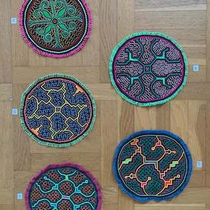 Könnte beinhalten: Fünf runde, bestickte Wandbehänge mit geometrischen Mustern in leuchtenden Farben, darunter Grün, Pink, Gelb und Orange, auf einem Holzgrund.