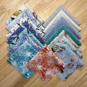 May include: A set of 12 fabric squares featuring various sea-themed patterns, including seashells, coral, and seabirds. The fabric squares are arranged in a fan shape on a wooden surface.