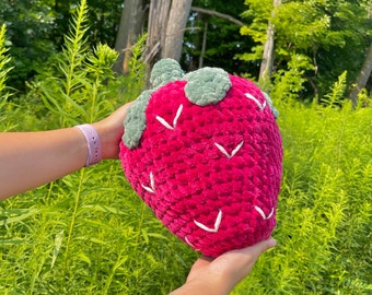 Peut inclure: Un coussin en forme de fraise en peluche de couleur rose foncé avec des coutures blanches. Le coussin a des feuilles en feutre vert sur le dessus.