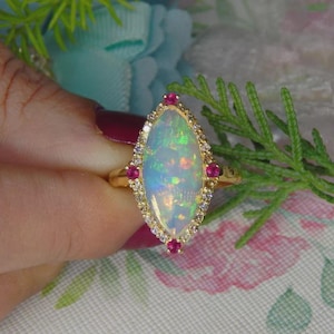 May include: A gold ring with an oval opal gemstone, displaying a play of colours. The opal is framed by small diamonds and red gemstones. The ring is set against a floral backdrop.