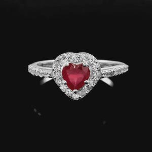 May include: A silver ring with a heart-shaped, deep red gemstone at the center, surrounded by a halo of smaller, clear stones. The band is also adorned with clear stones, creating a sparkling effect against a black background.