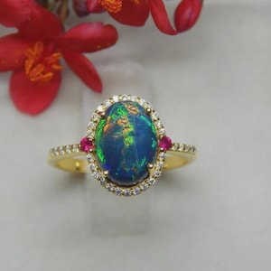 May include: A gold ring with an oval opal displaying blue, green, and yellow colours. The opal is encircled by small, clear gemstones and flanked by two red gemstones. The ring is set against a soft, white background. The ring's design is elegant.