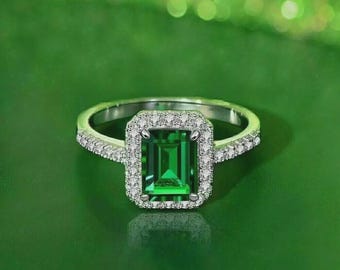 Puede incluir: Un anillo de plata con una gema verde rectangular con talla esmeralda, rodeada por un halo de piedras pequeñas y claras. La banda del anillo también está adornada con pequeñas piedras claras, creando un efecto brillante. El anillo está sobre un fondo verde.