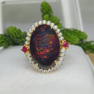 May include: A gold ring featuring a large, oval black opal with vibrant red and orange flashes. The opal is surrounded by a halo of small, clear diamonds and accented with two square-cut rubies. The ring is set on a white surface.