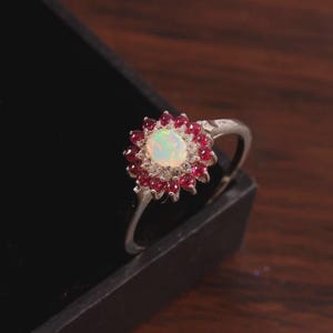 Op de afbeelding: Zilveren ring met een centrale opaalsteen, omringd door kleine rode edelstenen. Het ontwerp van de ring lijkt op een bloem, met de opaal in het midden en de rode stenen als bloemblaadjes. De ring staat tegen een donkere achtergrond.