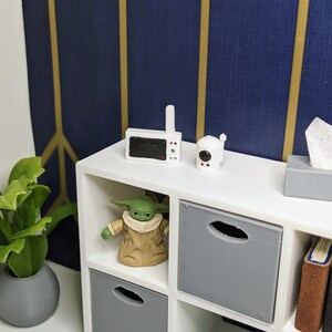 May include: A miniature white cube shelf unit with gray storage bins, holding a Grogu figurine, a small green frog, and books. A baby monitor and camera sit on top. A small plant in a gray pot is on the left.