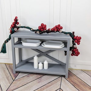 May include: A miniature gray shelf unit with two levels, holding white dishes and bottles. A decorative garland with red berries and clear beads adorns the top. The shelf is set against a white background and wooden floor.