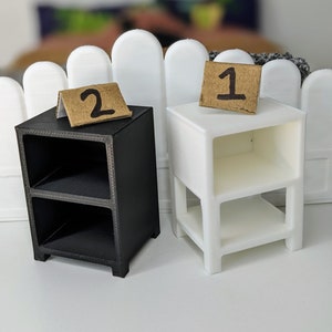 May include: Two miniature nightstands, one black and one white, each with two shelves. The black nightstand has a small cardboard sign with the number "2" on it, and the white nightstand has a sign with the number "1". The nightstands are in front of a white picket fence.
