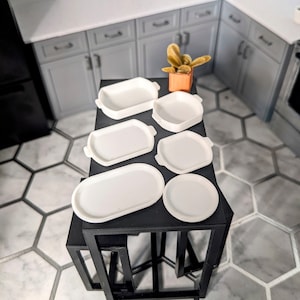 May include: A collection of six white miniature serving trays and plates, arranged on a black table. The trays vary in shape, including rectangular, oval, and round. The scene is set in a miniature kitchen with gray cabinets and a hexagonal tile floor.