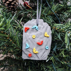 May include: A gray, rock-shaped Christmas ornament with colorful climbing holds. The ornament features red, yellow, blue, and orange holds, and is suspended by a gray ribbon. The ornament is displayed on a Christmas tree.