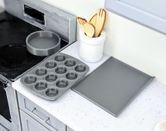 May include: A miniature kitchen scene featuring a gray muffin tin, a round baking pan, and a textured baking sheet. Wooden utensils rest in a white ceramic pot. The scene is set on a gray countertop with a gray and white speckled pattern.