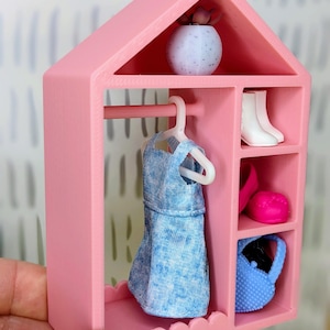 Puede incluir: Un armario rosa en miniatura para mu&ntilde;ecas con un vestido azul en una percha, botas blancas, un sombrero rosa y un bolso azul. Una peque&ntilde;a maceta moteada blanca y azul con una planta encima.