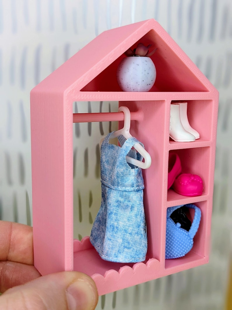 Puede incluir: Un armario rosa en miniatura para mu&ntilde;ecas con un vestido azul en una percha, botas blancas, un sombrero rosa y un bolso azul. Una peque&ntilde;a maceta moteada blanca y azul con una planta encima.