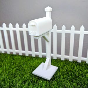 May include: A white mailbox with the word "MAIL" embossed on the front, mounted on a post. The mailbox is set in front of a white picket fence and green grass.