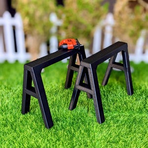 May include: Two black sawhorse sawhorses with an orange and black toy chainsaw on top, set against a backdrop of green grass and a white picket fence. The sawhorses are made of wood and have an A-frame design.