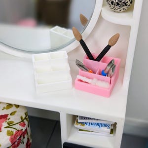 May include: A miniature white vanity with a round mirror, a pink organizer holding makeup brushes and tools, and a white organizer. A small stack of magazines is visible in a lower compartment. A decorative vase with a yellow flower sits on a shelf.