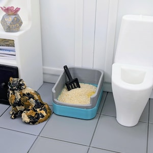 May include: A miniature cat figurine with a calico pattern sits next to a blue and gray cat litter box with a black scoop. A white toilet is in the background, and the scene is set on gray tile flooring.