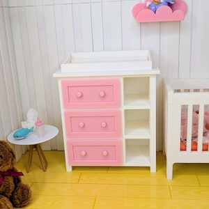 May include: A miniature nursery scene featuring a white and pink changing table with three drawers. A small white crib is to the right. A pink cloud shelf with a unicorn figurine is mounted on the wall. A teddy bear and small table are in the foreground.
