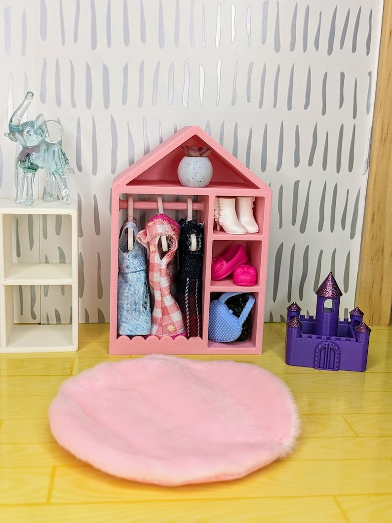 Puede incluir: Un armario rosa en miniatura para casa de mu&ntilde;ecas con ropa de mu&ntilde;ecas colgando, zapatos y un bolso azul. Una alfombra rosa est&aacute; colocada frente al armario. Un peque&ntilde;o castillo morado y un elefante de cristal tambi&eacute;n est&aacute;n en la escena.