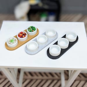 May include: Miniature serving trays with three white bowls each. One tray is tan, one is gray, and one is black. The bowls on the tan tray contain colorful food items. The trays are on a white table.
