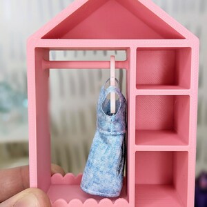 Puede incluir: Un armario rosa en miniatura para mu&ntilde;ecas con un vestido vaquero azul colgado dentro. El armario tiene forma de casa, una barra para colgar ropa y estantes. El armario es sostenido por una mano.