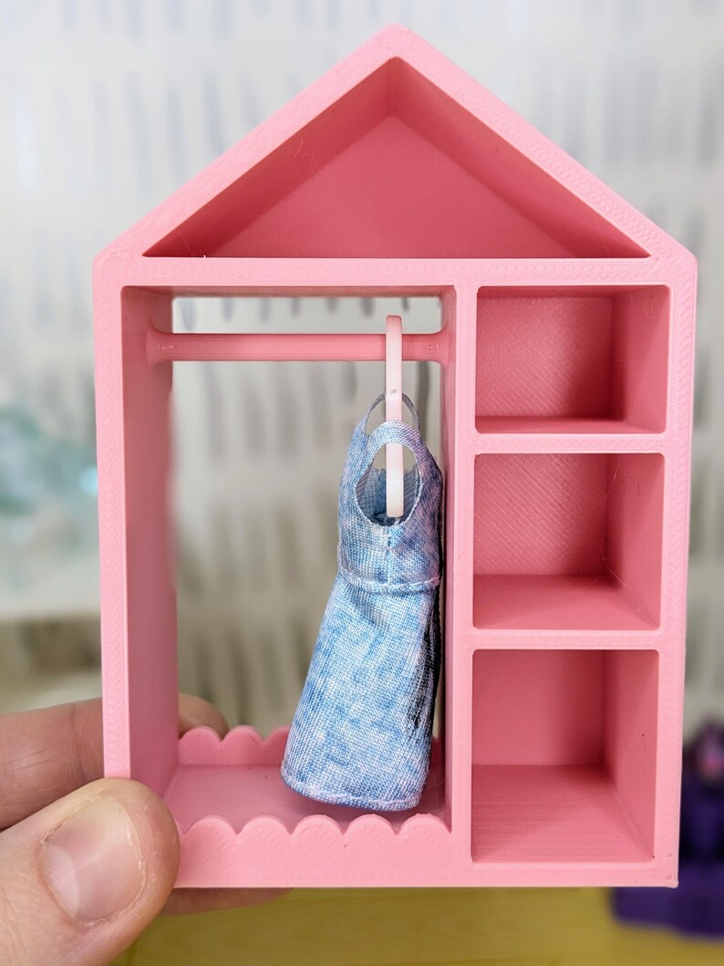 Puede incluir: Un armario rosa en miniatura para mu&ntilde;ecas con un vestido vaquero azul colgado dentro. El armario tiene forma de casa, una barra para colgar ropa y estantes. El armario es sostenido por una mano.