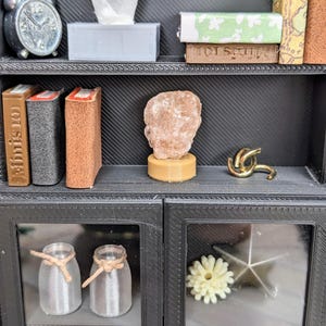 Puede incluir: Peque&ntilde;a estanter&iacute;a negra con varios objetos decorativos. Incluye un despertador plateado, una caja de pa&ntilde;uelos, libros, una escultura de roca, un nudo dorado, dos tarros de cristal con cuerda y una flor.