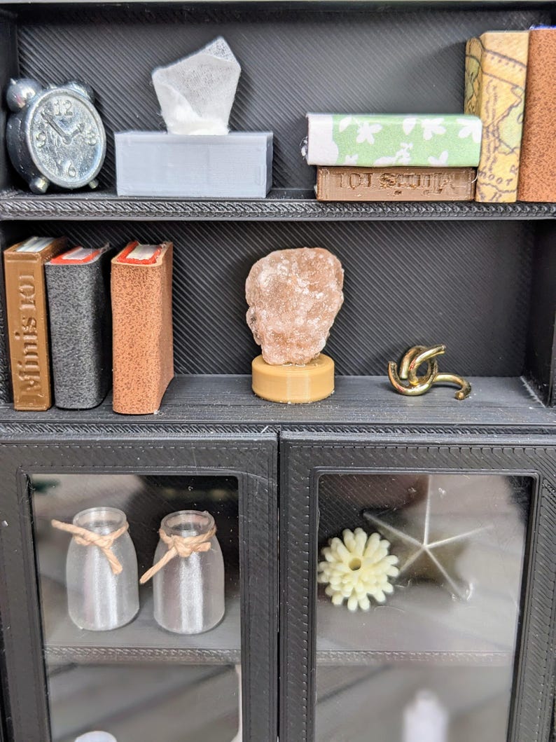 Puede incluir: Peque&ntilde;a estanter&iacute;a negra con varios objetos decorativos. Incluye un despertador plateado, una caja de pa&ntilde;uelos, libros, una escultura de roca, un nudo dorado, dos tarros de cristal con cuerda y una flor.