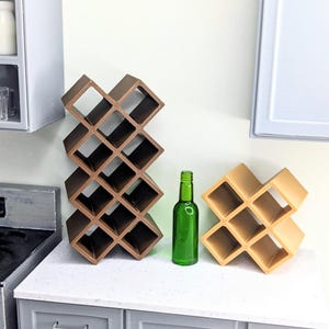 May include: Miniature brown and tan wine racks with a green glass bottle. The wine racks are designed with a grid pattern, and the bottle is empty. The items are on a white countertop with gray cabinets.