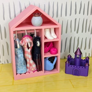Puede incluir: Un armario rosa para mu&ntilde;ecas con ropa de mu&ntilde;ecas colgada, botas blancas, zapatos rosas y un bolso azul. Un peque&ntilde;o castillo morado con un techo brillante est&aacute; al lado del armario. Una peque&ntilde;a planta decorativa est&aacute; encima del armario.
