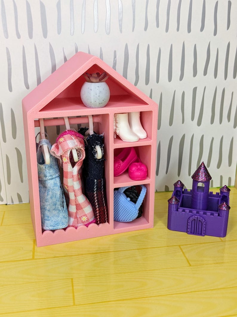 Puede incluir: Un armario rosa para mu&ntilde;ecas con ropa de mu&ntilde;ecas colgada, botas blancas, zapatos rosas y un bolso azul. Un peque&ntilde;o castillo morado con un techo brillante est&aacute; al lado del armario. Una peque&ntilde;a planta decorativa est&aacute; encima del armario.